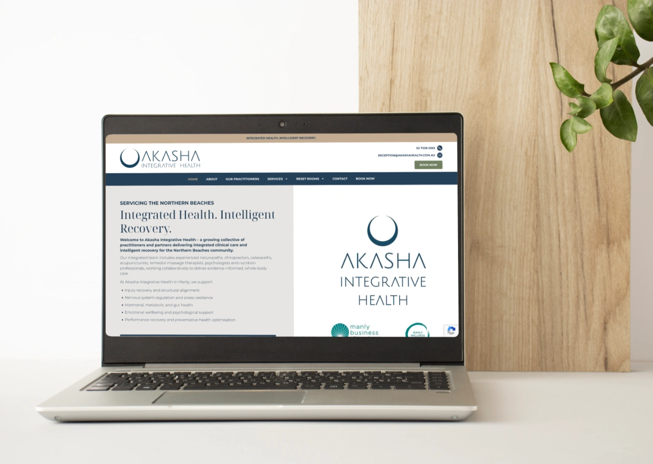 Laptop on a white desk displaying the Akasha Integrative Health website with its blue logo on screen.