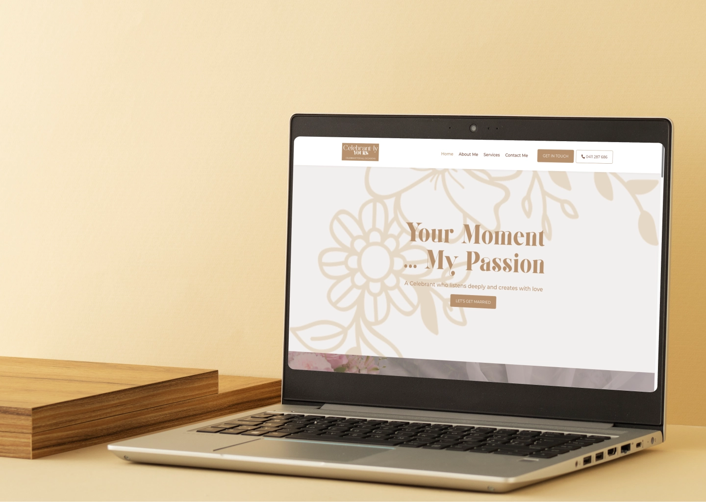 Laptop on a wooden desk displaying a wedding service website with floral background and tan text 'Your Moment ... My Passion'
